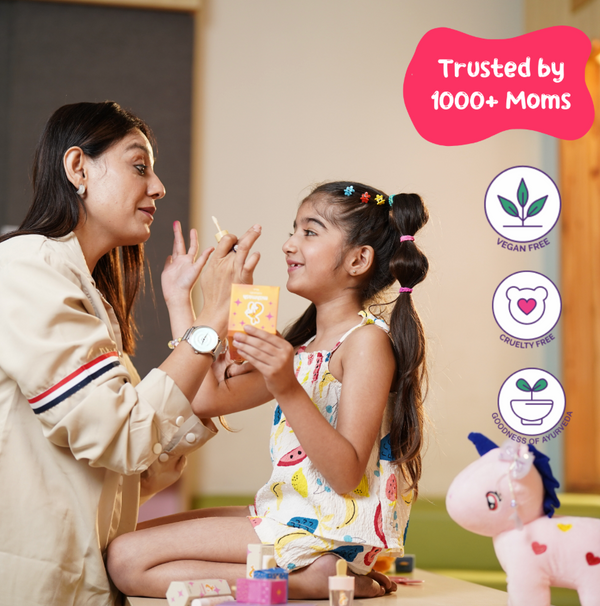A mother and daughter smiling while playing with kid-friendly mishmash makeup products. The daughter is wearing a colorful dress with fruit prints, and the mother is applying makeup on her face. Text on the image reads 'Trusted by 1000+ Moms' with icons for vegan free, cruelty free, and goodness of Ayurveda.